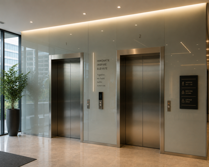 Modern elevator lobby with two stainless steel elevators, glass walls, warm recessed lighting, a potted plant, and a black directional sign on the right.